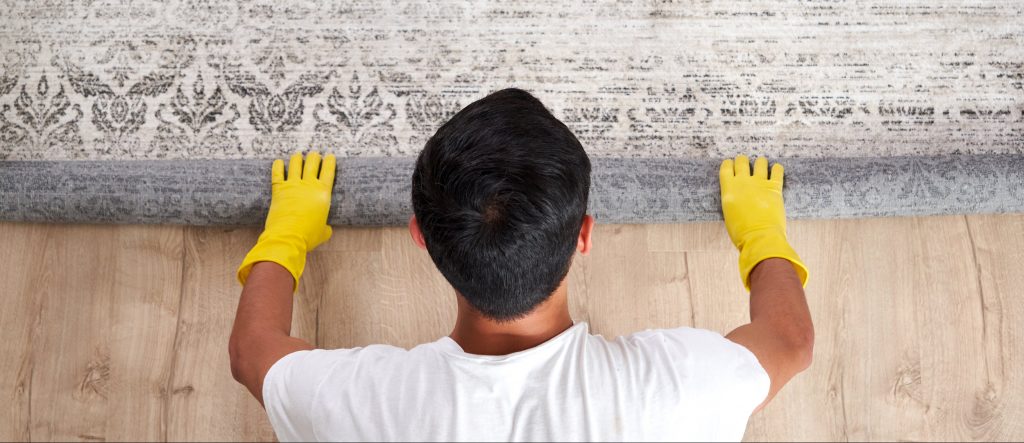 Mann rollt einen grauen Teppich zusammen und trägt dabei gelbe Gummi-Handschuhe.